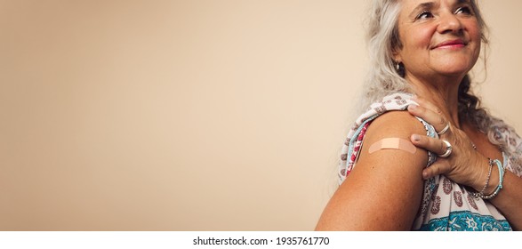 Close Up Of A Senior Woman Arm With A Bandage After Getting Vaccine Shot. Woman Getting A Flu Shot On Her Arm Standing Against Brown Background.