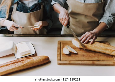 Close Up Of Senior Woman Hands Cutting Baguette. Granddaughter Hands Holding Knife With Butter. Making Sandwich. Grandma And Granddaughter Cooking Food. Healthy Lifestyle. Cooking At Home.