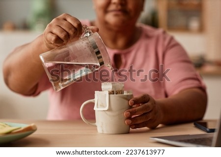 Close up of senior woman making tea in cozy home kitchen, holding glass kettle and pouring water, copy space