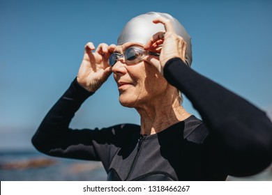 Close Up Of A Senior Woman In Swim Wear Standing Near The Sea. Female Swimmer Standing On Beach Wearing Her Swimming Cap And Goggles.