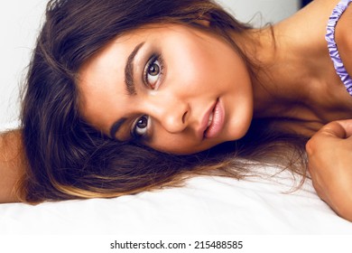 Close Up Sensual Portrait Of Beautiful Girl With Perfect Bronze Skin Big Brown Eyes And Full Lips Laying At Bed Relaxing And Enjoy Morning Time, Have Natural Glow Make Up And  Brunette Long Hairs.