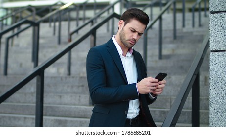 Close Up Serious Businessman Standing With Phone In Hand At Street. Disappointed Man Typing On Phone Outdoors At Stairs. Elegant Business Man Using Mobile Phone In Suit Outside.