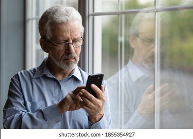 Close up serious mature man wearing glasses using phone, standing near window at home, lonely grandfather waiting for call from relatives or grandchildren, looking at smartphone screen - Powered by Shutterstock