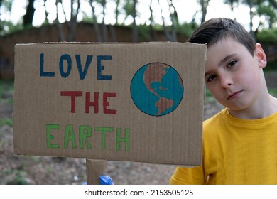 A Close Up Serious Young Boy Wearing A Yellow T-shirt And Blue Gloves Holding A Cardboard Sign Drawn By Hand With The Symbol Of A Terrestrial Globe And On Which He Puts An Ecological Awareness Message