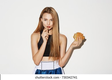 Close up of sexy blonde female model in sport wear eating free potato. holding burger in hand, looking in camera with flirty expression.