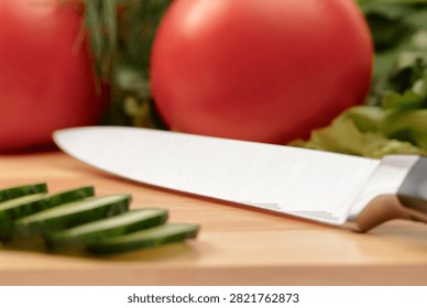 Close up of sharp chef knife beside sliced cucumber, tomato and fresh greens on cutting board.