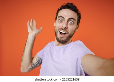 Close up of shocked young bearded man 20s in casual violet t-shirt standing doing selfie shot on mobile phone spreading hands isolated on bright orange background studio portrait Tattoo translate fun
