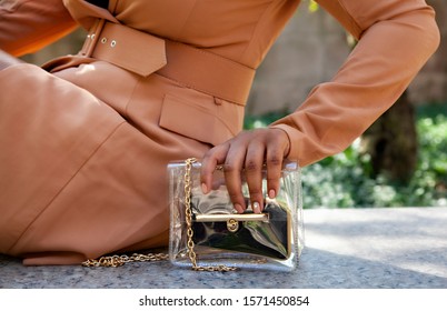 Close Up Shop Of A African American Black Woman Hands Holding A Clear Hand Bag By A Water Fountain In Chicago.  She's Wearing A Fashionable Brown Trench Coat.