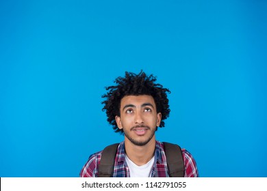 Close up short of a young man wearing a casual outfit and carry a backpack, looking up, standing on a blue background. - Powered by Shutterstock