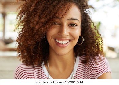 Close up shot of adorable African American woman has broad smile, wears striped t shirt, being in good mood, rests in cafeteria with best friends. Smiling dark skinned young female poses indoor