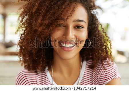 Close up shot of adorable African American woman has broad smile, wears striped t shirt, being in good mood, rests in cafeteria with best friends. Smiling dark skinned young female poses indoor