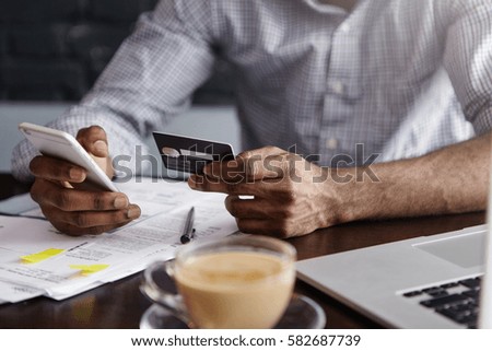 Close up shot of African man's hands holding mobile and credit card. Dark-skinned businessman checking account balance, using online banking application on his cell phone, drinking coffee at cafe