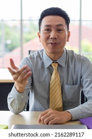 Close Up Shot Of Asia Short Black Hair Male Handsome Success Businessman Smile And Sitting Alone Behind Working Office Desk And Using Hand In Explain And Describe Gesture.