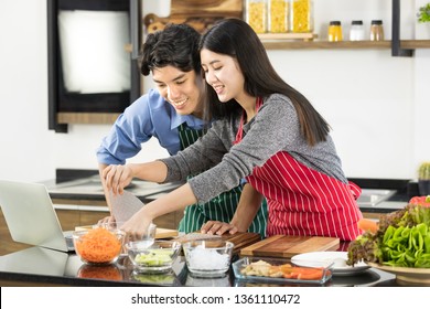 Close Up Shot Of Asian Couple In Apron Standing And Using Notebook To Find Recipe From Internet, Vegetables On Counter, In Kitchen, Concept For Foodie Lover.