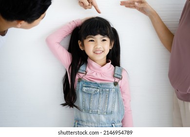 Close Up Shot Of Asian Small Cute Pigtail Hair Style Preschooler Child Daughter In Jeans Overalls Outfit Standing Smiling Leaning At Wall While Father Using Hand Measuring Checking Growth Rate Height.