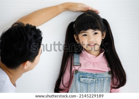 Close up shot of Asian small cute pigtail hair style preschooler child daughter in jeans overalls outfit standing smiling leaning at wall while father using hand measuring checking growth rate height.