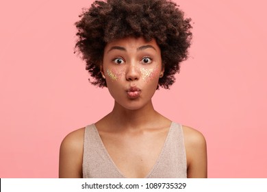 Close up shot of attractive African female keeps lips round, has curly dark hair, lovely face decorated with shining glitter, flirts with handsome guy indoor. People, beauty and cosmetics concept