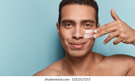 Close up shot of attractive man with healthy skin, applies cream for anti wrinkle or anti aging, cares of body, poses naked or shirtless, isolated over blue background with free space for your text