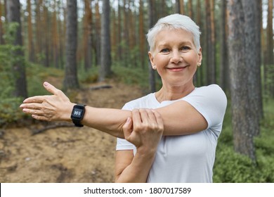 Close Up Shot Of Attractive Short Haired Retired Female Wearing Whit T-shirt And Smart Watch On Her Wrist To Track Progress During Running, Stretching Arm, Preparing Body For Cardio Exercise