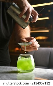 Close Up Shot Of Bartender Hand Pouring Basil Cocktail In A Glass From Shaker. Barman Preparing Classic Modern Basil Smash Cocktail.