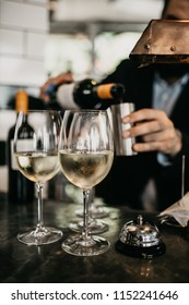 A Close Up Shot Of A Bartender Measuring And Pouring White Wine Into The Wine Glasses On The Bar.Selective Focus On The Full Glasses Of Wine. Hospitality And Restaurant Concept.