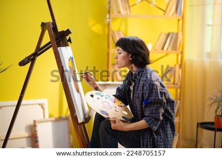 A close up shot of the beautiful and alluring woman painting something modern and majestic onto her white canvas in her very modern and bright art studio