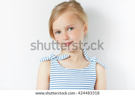Close up shot of beautiful blonde Caucasian little girl wearing striped dress, looking at the camera with adorable smile, posing against white copy space concrete wall. Happy childhood concept