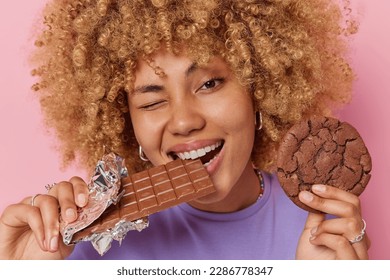 Close up shot of beautiful woman with curly hair bites bar of chocolate holds cookie winks eye has perfect white teeth has addiction to sugar isolated over pink background. Sweet tooth concept