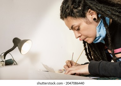 Close Up Shot Of Beautiful Young White Girl Hispanic Latin Art Director, With Dreadlocks And Good Style, In Her Production Studio Concentrating On Her Work Table Lit By A Lamp, Tracing An Onion Paper.