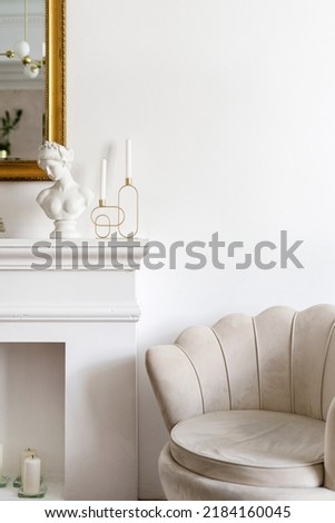 Close up shot of beige soft chair in classic style near decorative fireplace. Bust figurine, metal candlestick with candles and mirror with golden frame in living room with elegant interior design