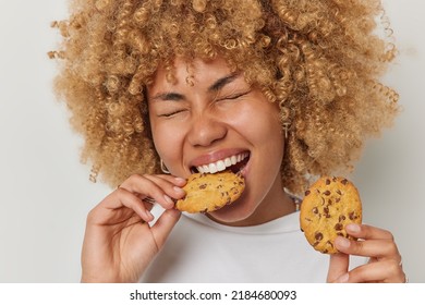Close up shot of blonde curly haired woman eats cookies with chocolate bites deicious snack keeps eyes closed has sweet tooth dressed casually isolated over white background. Unhealthy eating