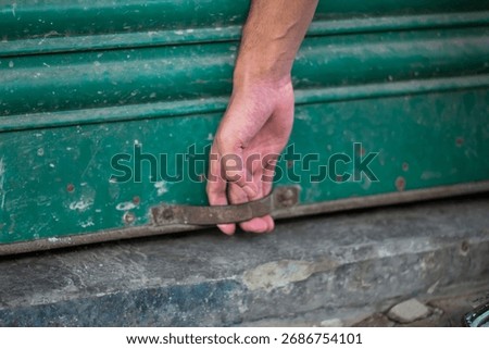A close up shot captures a person's hand gripping the metal handle of a distressed, green rolling door, highlighting the texture and wear of the surface.