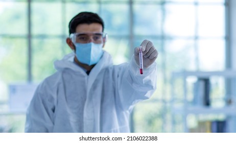 Close Up Shot Of Caucasian Professional Bearded Male Scientist In White Ppe Lab Coat Safety Goggles And Face Mask Holding Looking At Blood Sample Test Tube In Hand With Laboratory Science Equipment.