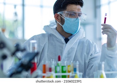 Close Up Shot Of Caucasian Professional Bearded Male Scientist In White Ppe Lab Coat Safety Goggles And Face Mask Holding Looking At Blood Sample Test Tube In Hand With Laboratory Science Equipment.