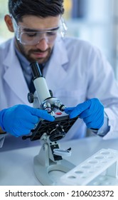 Close Up Shot Of Caucasian Professional Bearded Male Scientist In White Ppe Lab Coat Safety Goggles And Face Mask Holding Looking At Blood Sample Test Tube In Hand With Laboratory Science Equipment.