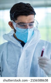 Close Up Shot Of Caucasian Professional Bearded Male Scientist In White Ppe Lab Coat Safety Goggles And Face Mask Holding Looking At Blood Sample Test Tube In Hand With Laboratory Science Equipment.