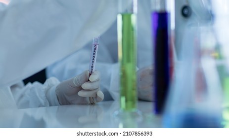 Close Up Shot Of Caucasian Professional Bearded Male Scientist In White Ppe Lab Coat Safety Goggles And Face Mask Holding Looking At Blood Sample Test Tube In Hand With Laboratory Science Equipment.