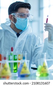 Close Up Shot Of Caucasian Professional Bearded Male Scientist In White Ppe Lab Coat Safety Goggles And Face Mask Holding Looking At Blood Sample Test Tube In Hand With Laboratory Science Equipment.