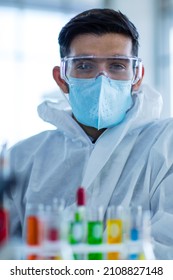 Close Up Shot Of Caucasian Professional Bearded Male Scientist In White Ppe Lab Coat Safety Goggles And Face Mask Holding Looking At Blood Sample Test Tube In Hand With Laboratory Science Equipment.