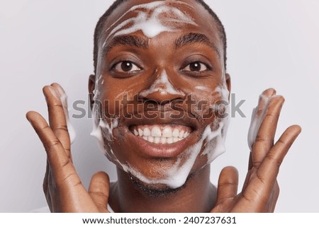 Close up shot of cheerful dark skinned man applies cleansing gel on face has fun whille washing cheeks involved in skin care routine smiles toorhily keeps hands raised up isolated on white background