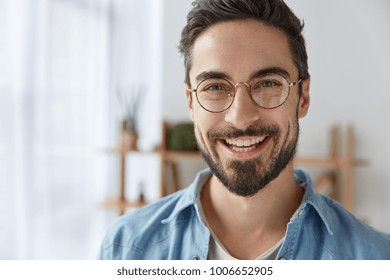 Close up shot of cheerful satisfied attractive male with stubble, has broad smile, wears round spectacles, rejoices success at work, stands against cozy interior. Fashionable designer glad be praised - Shutterstock ID 1006652905