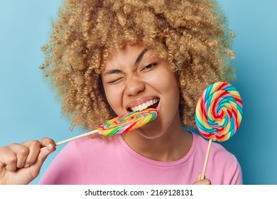 Close Up Shot Of Cheerful Teenage Girl Bites Sweet Multicolored Lollipop Enjoys Eating Sugary Food Winks Eyes Dressed In Casual T Shirt Isolated Over Blue Background. Woman With Caramel Candy