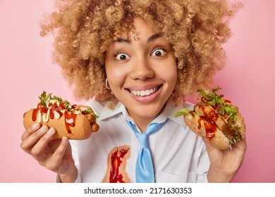 Close Up Shot Of Cheerful Woman Looks With Big Surprised At Camera Smiles Broadly Holds Fast Food Enjoys Eating Tasty Sandwich And Hot Dog Dressed In Formal Clothes Smeared As Being Careless