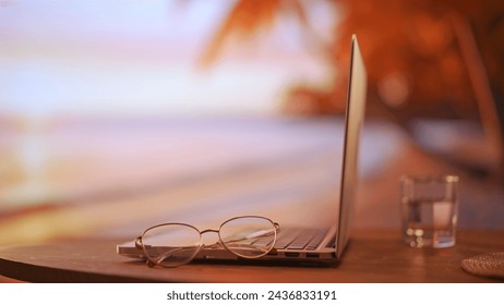 Close up shot of computer at the table. Closed laptop on the table with glasses and glass of water against tropical beach seaside background.