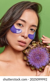 Close up shot of confident serious Asian woman with dark hair healty skin uses natural cosmetic products made of flowers applies blue hydrogel patches under eyes to reduce puffiness and moisturize