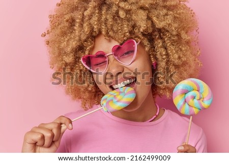 Close up shot of curly haired young woman bites multicolored lollipops has sweet tooth enjoys favorite dessert wears heart shaped sunglasses and casual t shirt isolated over pink background.