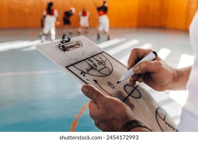 Close shot of an elderly caucasian male basketball coach drawing his strategy on a coach clipboard with team in background during a practice in a sports hall. Copy space. - Powered by Shutterstock