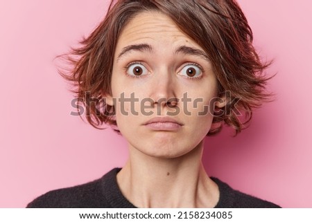 Close up shot of embarrassed woman with hairstyle has widely opened eyes from fright feels worried isolated over pink background reacts to scary news has emotional stress. Human reactions concept