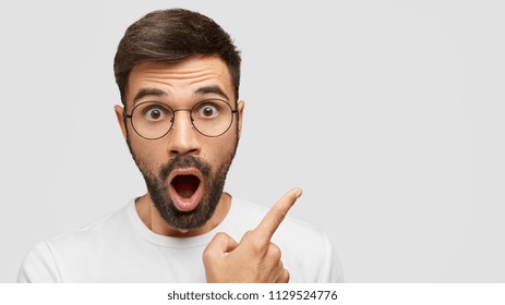 Close up shot of emotive shocked bearded young male opens mouth widely, stare at camera, points upwards aside at blank space, says: Wow, it`s something unbelievable! People and emotions concept - Shutterstock ID 1129524776