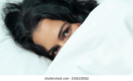 Close Up Shot Of An Ethnic Female Looking At The Camera, While Hiding Her Face Under The Blanket, Peeking Through. Beautiful Woman Waking Up In A Fancy Hotel Room To Celebrate Valentines Day.